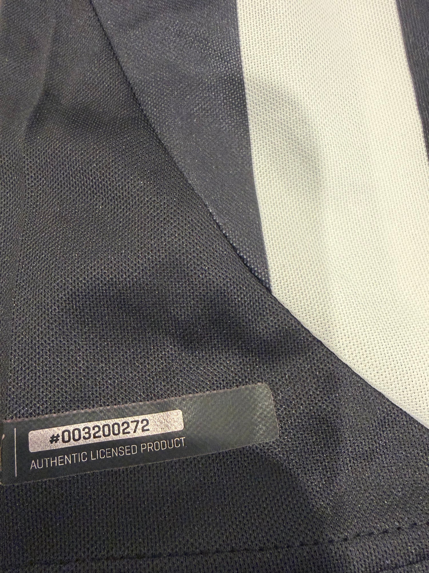 Close-up of a dark navy athletic garment with a light gray vertical stripe and a transparent authenticity label reading 'AUTHENTIC LICENSED PRODUCT' and '#00320072'.